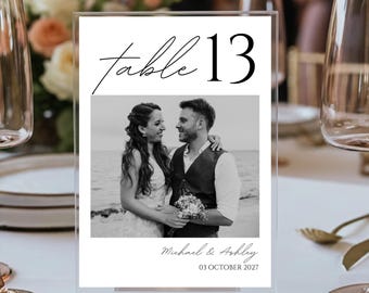May include: Clear acrylic table number 13 with a black and white photo of a couple. The photo is below the words "table 13" in a script font. The names "Michael & Ashley" and the date "03 OCTOBER 2027" are below the photo.
