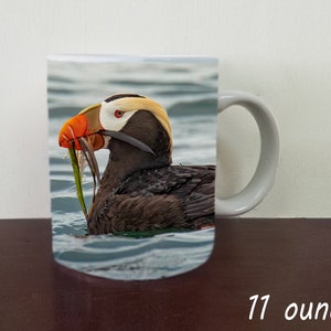 May include: A white ceramic mug with a colorful image of a puffin with an orange beak and yellow head swimming in the ocean. The mug is 11 ounces.