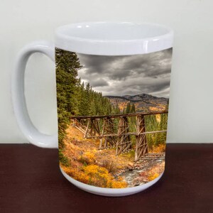 May include: White ceramic mug with a scenic image of a wooden trestle bridge in a mountainous landscape. The bridge is surrounded by trees and a river, with a cloudy sky above.