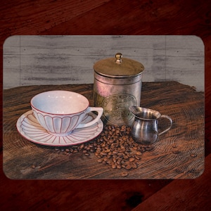 May include: A white teacup with a red rim sits on a saucer with a red and white floral design. The teacup is next to a silver creamer and a silver canister with the text "ORIGINAL COFFEE" on it. The items are on a wooden surface with coffee beans scattered around them.