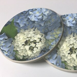 May include: Two round coasters with a floral design. The coasters feature a close-up of blue and white hydrangeas. The text "Agatha Christie" is visible on one coaster.