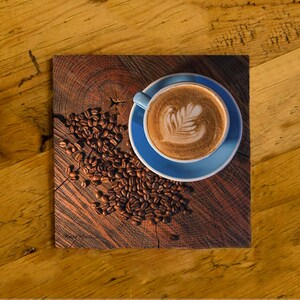 May include: A blue ceramic saucer with a white cup of coffee with latte art on top. The cup and saucer are on a wooden surface with coffee beans scattered around them.