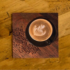 May include: A cup of coffee with latte art in a black mug on a dark wooden coaster. Coffee beans are scattered around the coaster. The coaster is on a wooden table.