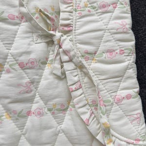 Könnte beinhalten: Weiß gesteppte Babydecke mit einem Blumenmuster in Rosa, Gelb und Grün. Die Decke hat ein Rautenmuster und einen gerüschten Rand. Ein Krawattenverschluss ist sichtbar.