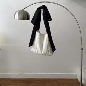May include: A white quilted tote bag with a black strap is suspended from a silver arc lamp. The bag has a soft, puffy appearance. The lamp has a round, brushed metal shade.