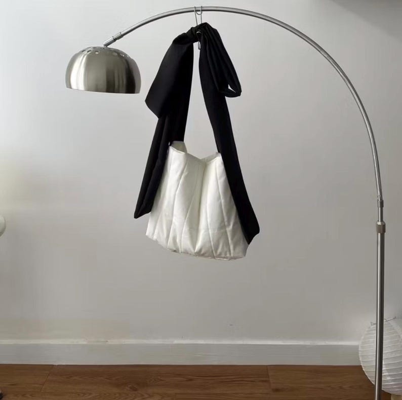 May include: A white quilted tote bag with a black strap is suspended from a silver arc lamp. The bag has a soft, puffy appearance. The lamp has a round, brushed metal shade.