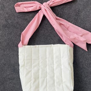 May include: A white quilted tote bag with a pink bow-shaped handle. The bag has a rectangular shape with vertical stitching. The pink handle is tied in a bow at the top.