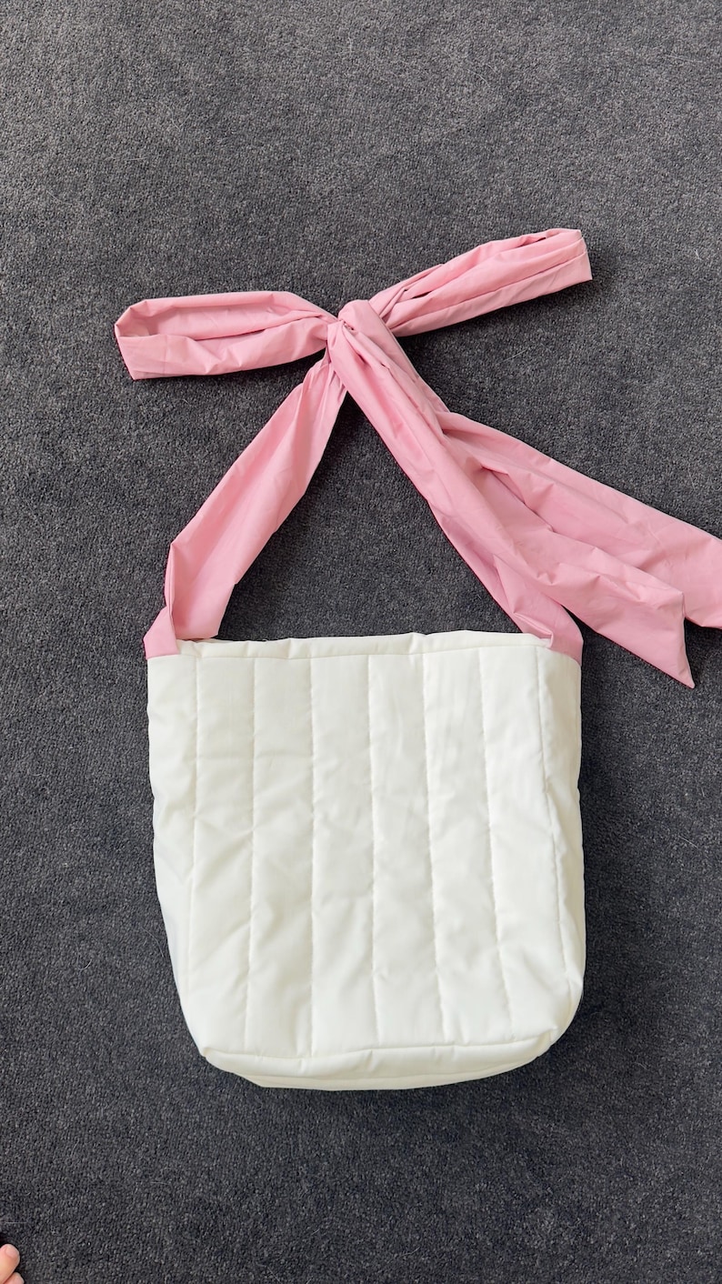 May include: A white quilted tote bag with a pink bow-shaped handle. The bag has a rectangular shape with vertical stitching. The pink handle is tied in a bow at the top.