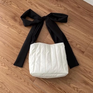 May include: A white, quilted tote bag with a large black bow detail. The bag has a rectangular shape and a padded texture. The black bow is positioned at the top, creating a striking contrast against the white bag. The bag is on a wooden surface.