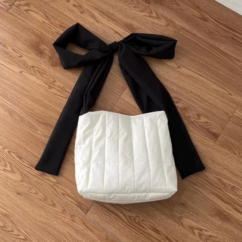 May include: A white, quilted tote bag with a large black bow detail. The bag has a rectangular shape and a padded texture. The black bow is positioned at the top, creating a striking contrast against the white bag. The bag is on a wooden surface.