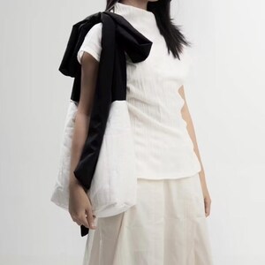 May include: A white, textured short-sleeve top and skirt. A black scarf is draped over the shoulder, and a white quilted tote bag is carried. The outfit is minimalist and modern, with a focus on texture and contrast.