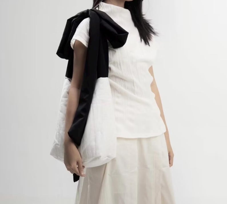 May include: A white, textured short-sleeve top and skirt. A black scarf is draped over the shoulder, and a white quilted tote bag is carried. The outfit is minimalist and modern, with a focus on texture and contrast.