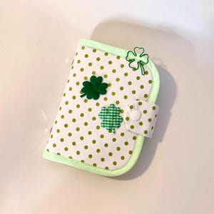 May include: A white and green polka-dot notebook cover with a pale green border. The cover features three shamrock embellishments: two solid green and one green gingham. A white button secures the flap.