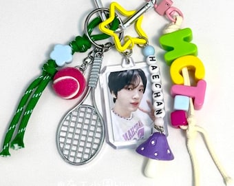 May include: A colorful keychain with a tennis racket, a tennis ball, a mushroom charm, a star charm, and a photo card with the name "HAECHAN" spelled out in beads. The keychain also has a green, white, and pink rope.