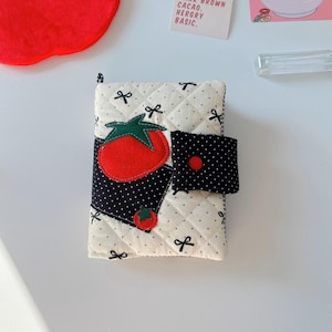Könnte beinhalten: Eine weiße gesteppte Stofftasche mit einer schwarzen und weißen Tupfenklappe. Die Tasche hat eine rote Tomatenapplikation mit grünen Blättern und einer schwarzen Schleife auf der Vorderseite. Die Tasche hat einen kleinen schwarzen Knopf auf der Klappe.