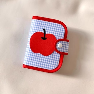 May include: A small blue and white gingham fabric wallet with a red apple applique. The wallet has a red trim and a white button closure.