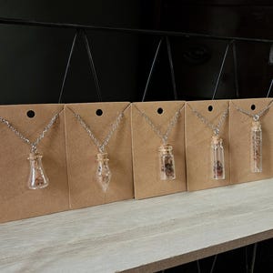 May include: Five silver-toned metal chain necklaces, each featuring a small, clear glass vial pendant sealed with a cork stopper. Inside each vial are small, dried botanical elements. The necklaces are displayed on individual brown cardstock tags.