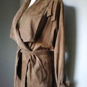 May include: A long brown suede trench coat with a wrap-style collar and a tie belt. The coat has long sleeves with a tie detail at the cuffs.