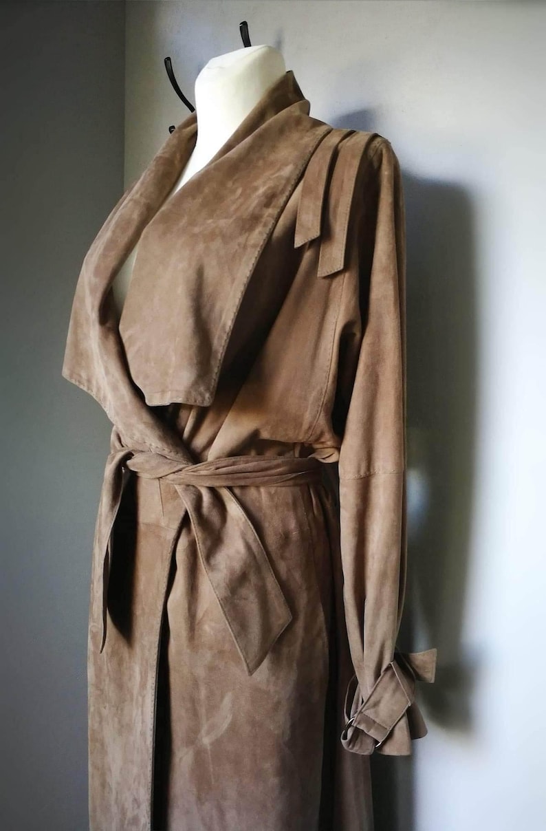 May include: A long brown suede trench coat with a wrap-style collar and a tie belt. The coat has long sleeves with a tie detail at the cuffs.