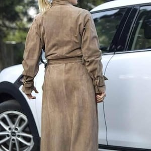 May include: A woman wearing a brown suede trench coat with a belt. She is standing next to a white car.