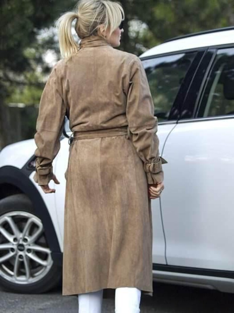May include: A woman wearing a brown suede trench coat with a belt. She is standing next to a white car.