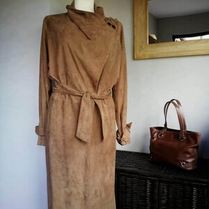May include: A long brown suede coat with a tie belt and a high collar. The coat is on a mannequin and is displayed against a white wall.