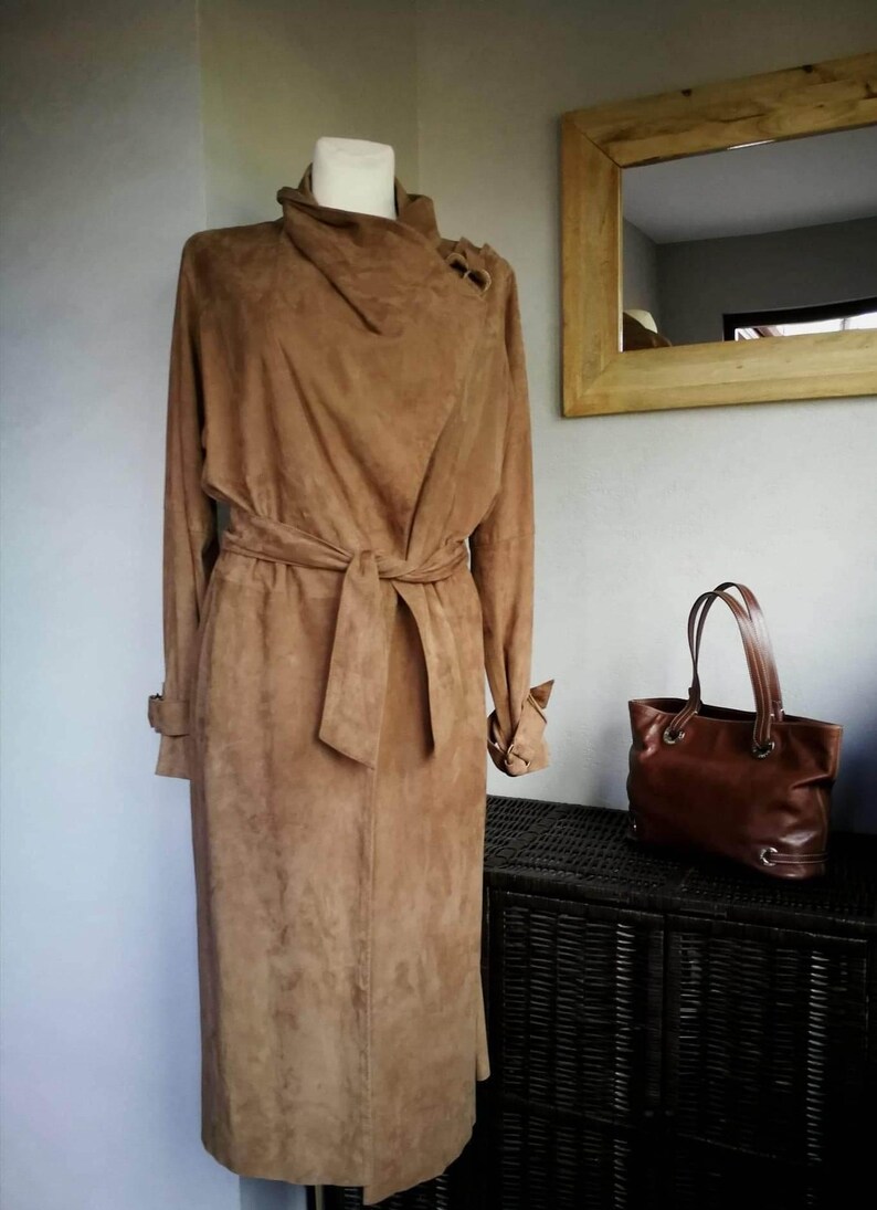 May include: A long brown suede coat with a tie belt and a high collar. The coat is on a mannequin and is displayed against a white wall.