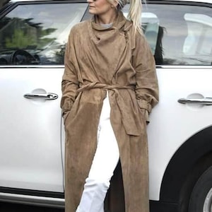 May include: A woman wearing a long brown suede coat with a belt, white pants, and sunglasses. She is standing next to a white car.