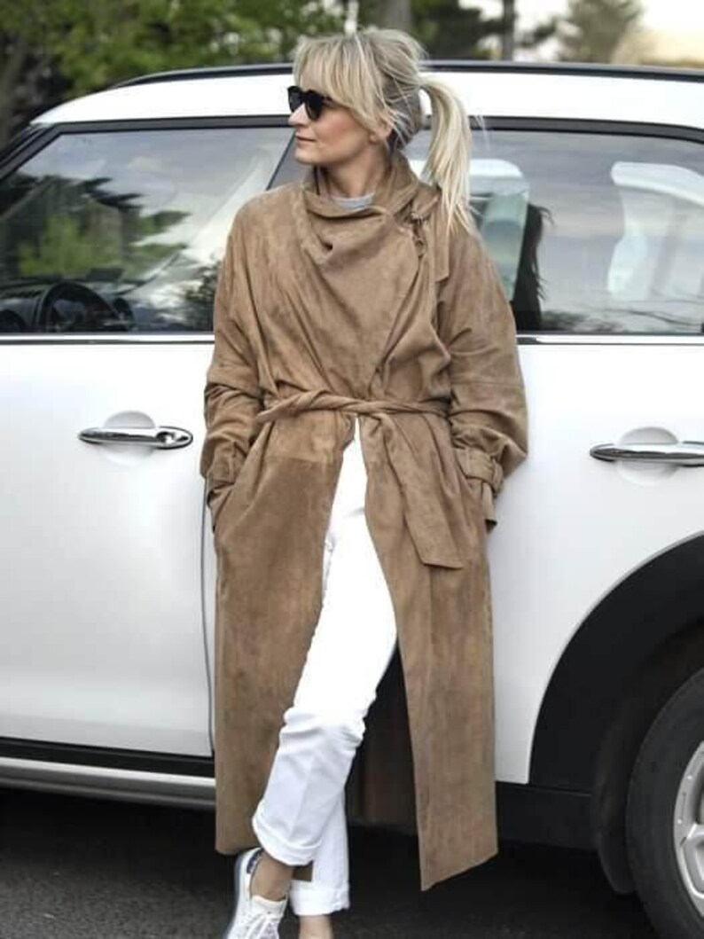 May include: A woman wearing a long brown suede coat with a belt, white pants, and sunglasses. She is standing next to a white car.