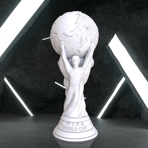 Puede incluir: Un trofeo blanco de la Copa Mundial de la FIFA. El trofeo presenta una figura sosteniendo un globo terráqueo. La base circular lleva la inscripción "FIFA WORLD CUP". El fondo es una superficie oscura y texturizada con iluminación lineal blanca.