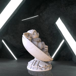 May include: A white ceramic figurine of a character from the Star Wars franchise, sitting in a pod-like chair. The figurine is on a white base with a textured surface.