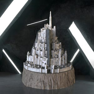 May include: A detailed white and grey scale model of a multi-tiered fortress city built into a steep mountain peak. The structure features numerous small buildings, a high central tower, and a fortified outer wall, all resting on a rocky base.