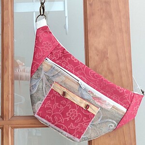 May include: A handmade fanny pack with a floral pattern in shades of red and gray. The bag has a white zipper, a small front pocket, and a cream-colored strap. The bag is hanging from a white hook on a wooden door.