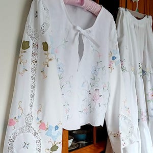 May include: White embroidered blouse and skirt set. The blouse features a round neckline with a tie closure and long sleeves with lace detailing. The set is adorned with floral embroidery in pastel colors, creating a delicate and feminine look.