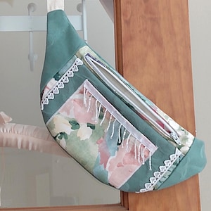 May include: A green fanny pack with a floral print and white lace trim. The bag has a zipper closure and a white strap.