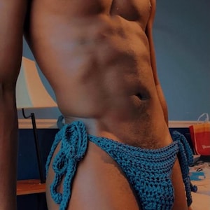 May include: A teal blue crochet bikini bottom featuring side ties with a textured knit pattern. The garment includes the text @skinnyguytry visible in the upper right corner of the frame. The piece is constructed with a visible yarn stitch.