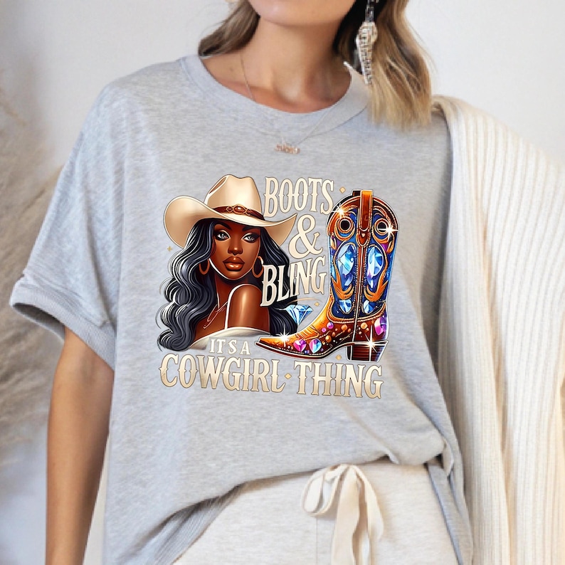 K&ouml;nnte beinhalten: Hellgraues T-Shirt mit einer Grafik einer Cowgirl mit Hut und einem verzierten Cowboy-Stiefel. Der Text auf dem Shirt lautet "Boots & Bling, It's a Cowgirl Thing."