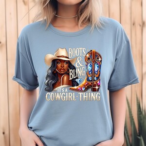 K&ouml;nnte beinhalten: Hellblaues T-Shirt mit einem Aufdruck einer Frau mit Cowboyhut und einem verzierten Cowboy-Stiefel. Der Text auf dem Shirt lautet "BOOTS & BLING" und "IT'S A COWGIRL THING."