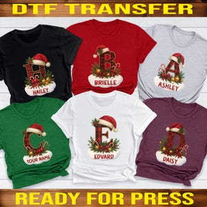 May include: DTF transfer designs featuring Christmas-themed graphics on various colored t-shirts. Each shirt displays a letter in a plaid pattern, a Santa hat, and a name. The text "DTF TRANSFER" and "READY FOR PRESS" are visible.