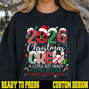 May include: A black crewneck sweatshirt featuring a graphic with the text 2026 CHRISTMAS CREW A LITTLE BIT CRAZY A LITTLE BIT LOUD A WHOLE LOTTA LOVE. The design includes red and green patterns, Santa hats, snowflakes, and candy canes.