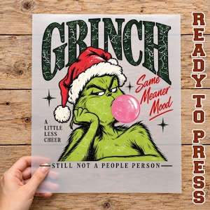 May include: A screen print transfer featuring a green Grinch character wearing a red Santa hat and blowing a pink bubble. Text includes GRINCH, A LITTLE LESS CHEER, Same Meaner Mood, and STILL NOT A PEOPLE PERSON in black and red lettering.