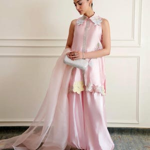 May include: A sleeveless light pink satin tunic with a collared neckline, featuring white floral embroidery on the collar and hem, paired with matching wide-leg satin trousers, a sheer pink organza dupatta, and a silver metallic rectangular clutch.