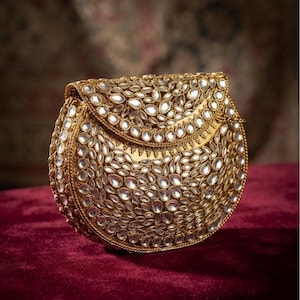 May include: A semi-circular gold clutch bag featuring an intricate surface covered in numerous teardrop-shaped white stones. The bag has a structured flap closure and a metallic gold finish, with the stones arranged in a dense, repeating pattern.