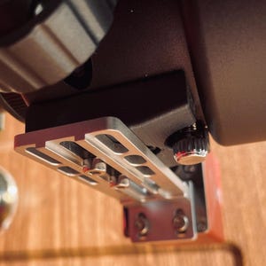 May include: Close-up of a black and silver camera accessory. The device has a silver metal plate with multiple slots and screws, attached to a black base. A red component is visible, along with a black knob.