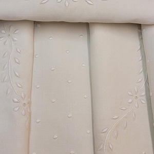 Puede incluir: Un tejido de color crema con un delicado bordado blanco de pequeñas flores y hojas dispuestas en patrones curvos. La tela está adornada con un sutil patrón de puntos en relieve y terminada con un borde festoneado en la parte inferior.