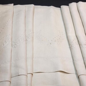 Puede incluir: Un mantel de color blanco roto plegado que presenta delicados patrones de coronas florales bordadas en blanco a lo largo de los bordes. La tela tiene una textura suave y está dispuesta en múltiples pliegues verticales.