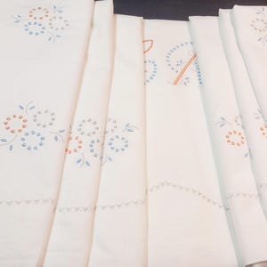 Puede incluir: Un juego de ropa de cama blanca con motivos florales bordados en hilo azul claro y naranja. La tela presenta un borde festoneado con costuras decorativas, dispuesta en capas plegadas para mostrar los patrones repetitivos.