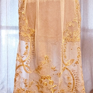 May include: A cream-colored skirt with intricate gold embroidery and sequin details. The skirt features a sheer overlay with floral and scroll patterns. A wide, bright yellow satin sash is at the waist. The skirt is likely for special occasions.