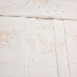Op de afbeelding: Een wit rechthoekig tafelkleed met lichtbruine geborduurde bloemmotieven en decoratieve geschulpte randen. De stof vertoont ingewikkelde borduurpatronen met delicaat stikwerk langs de randen en in de centrale delen van het textiel.