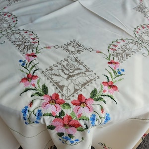 Peut inclure: Une nappe de couleur crème ornée de broderies au point de croix. Le motif comprend des formes géométriques marron et des motifs floraux dans des tons de rose, rouge et bleu, disposés en une bordure répétitive sur la surface du tissu.